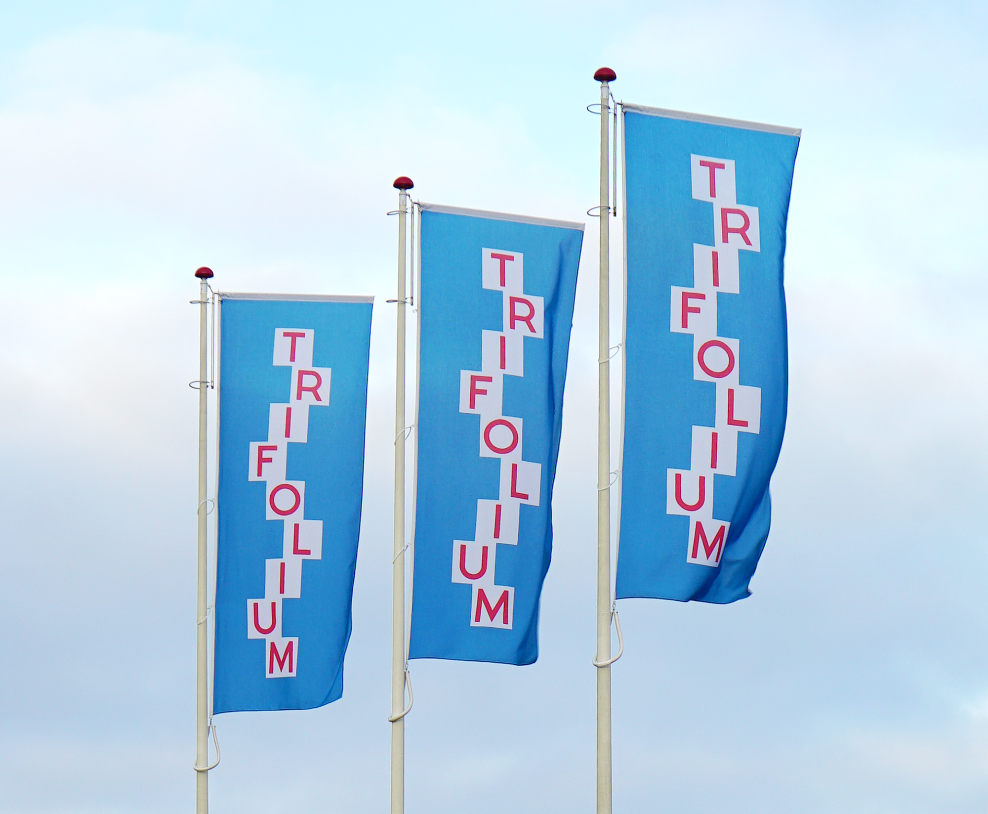 Three large flags with vertical Trifolium logo graphics on a cloudy sky